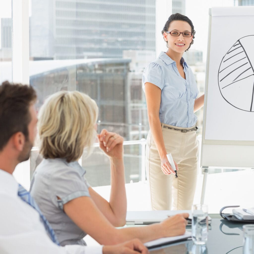 Mujer presentando un gráfico de pastel en una reunión de trabajo, con dos colegas sentados en la mesa, durante una presentación corporativa en oficina moderna